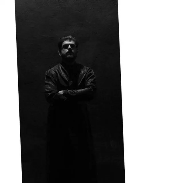 Ασπρόμαυρο πορτρέτο άνδρα με μακρύ σκούρο ένδυμα και σταυρωμένα χέρια σε στενό άνοιγμα.