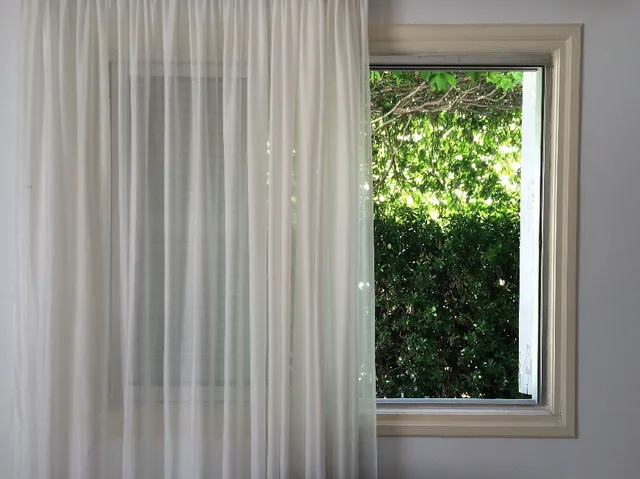 Λευκή διάφανη κουρτίνα δίπλα σε ανοιχτό παράθυρο με πράσινα φύλλα έξω.