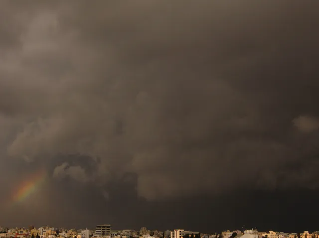 Τμήμα ουράνιου τόξου ξεπροβάλλει πάνω από την πόλη κάτω από βαριά σκοτεινά σύννεφα.