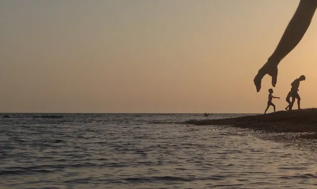 Ένα χέρι σε πρώτο πλάνο μοιάζει να αγγίζει δύο μακρινές ανθρώπινες σιλουέτες στην ακτή.
