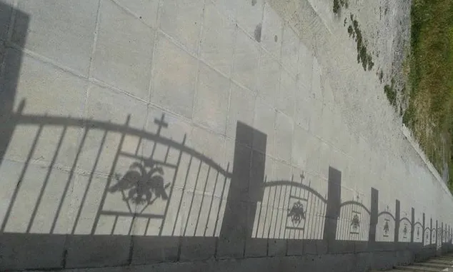 Σκιά περίτεχνου μεταλλικού κιγκλιδώματος πάνω σε ανοιχτόχρωμο πεζοδρόμιο.