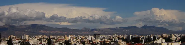 Πανοραμική άποψη της πόλης με βουνά στον ορίζοντα και μεγάλα λευκά σύννεφα.